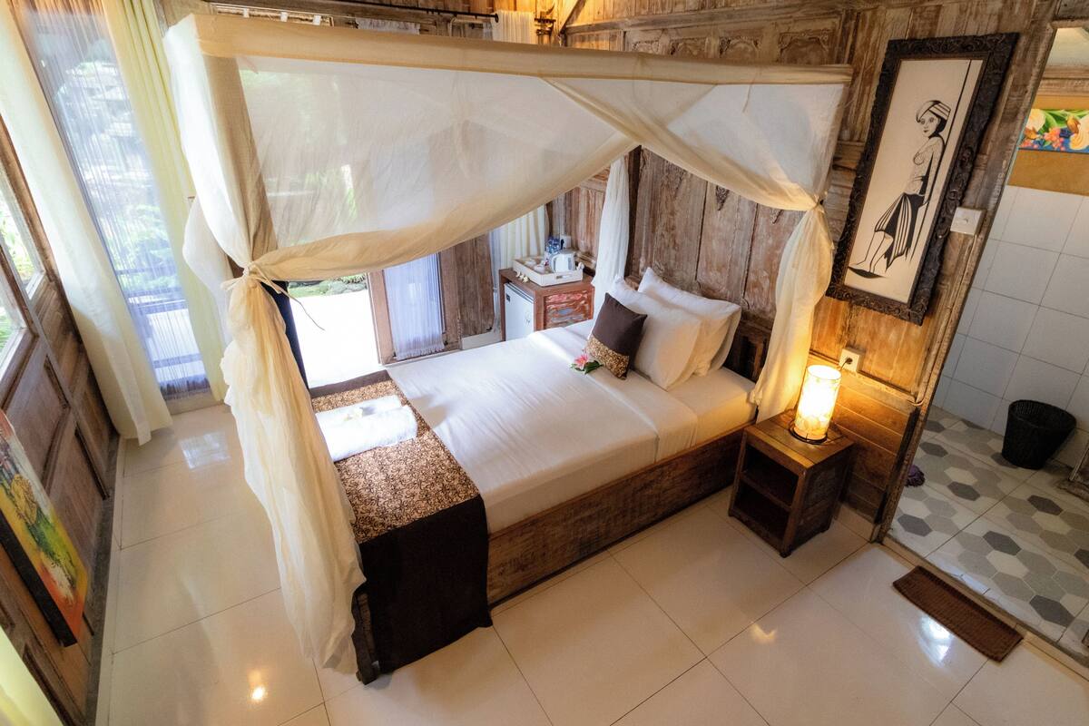 A room with a bed under a canopy, a bedside table with a lamp, a small wooden nightstand, and a framed picture on the wall.