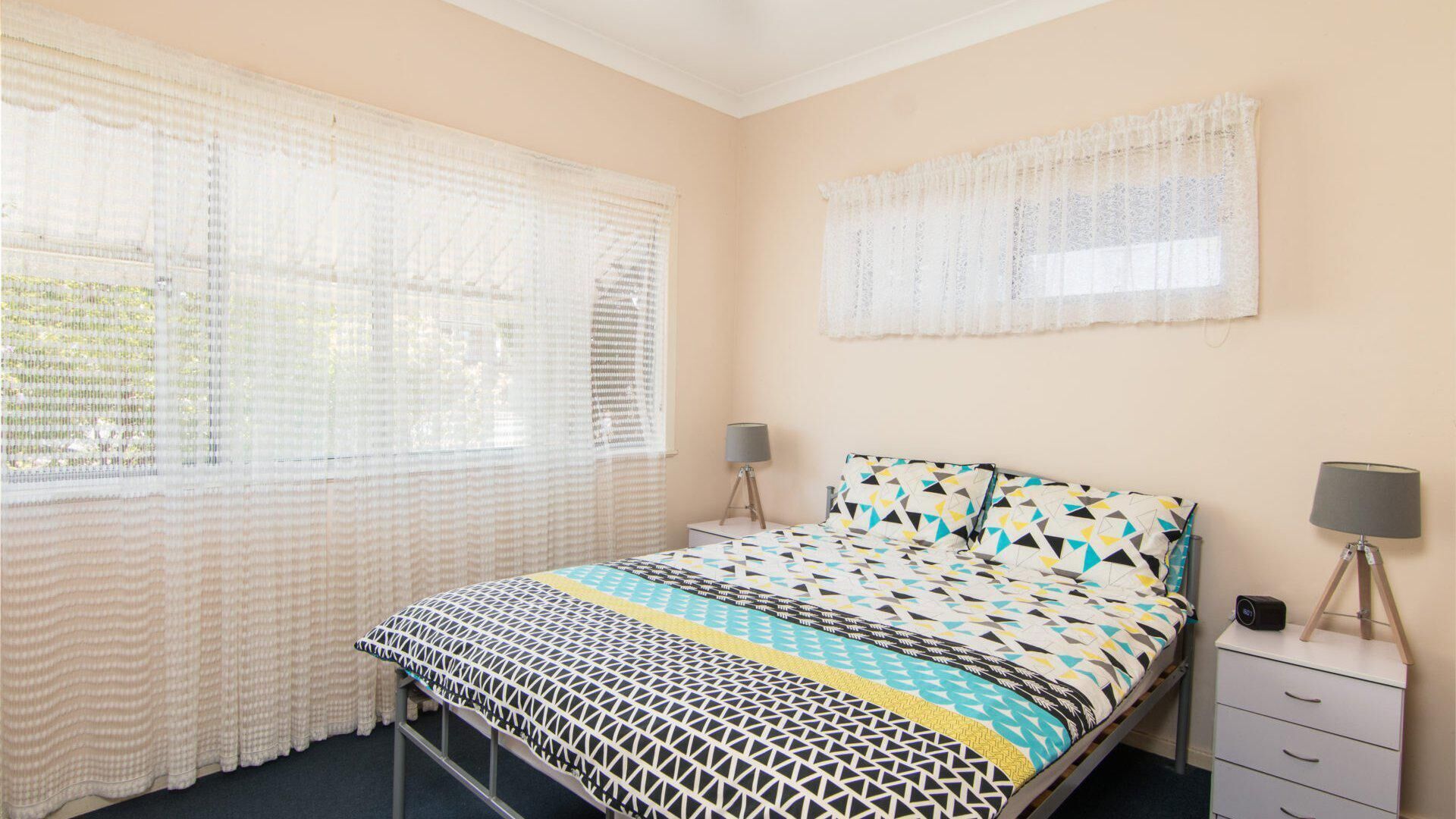 A bedroom with a bed, two bedside tables with lamps, a window with sheer curtains, and a wall-mounted light fixture.