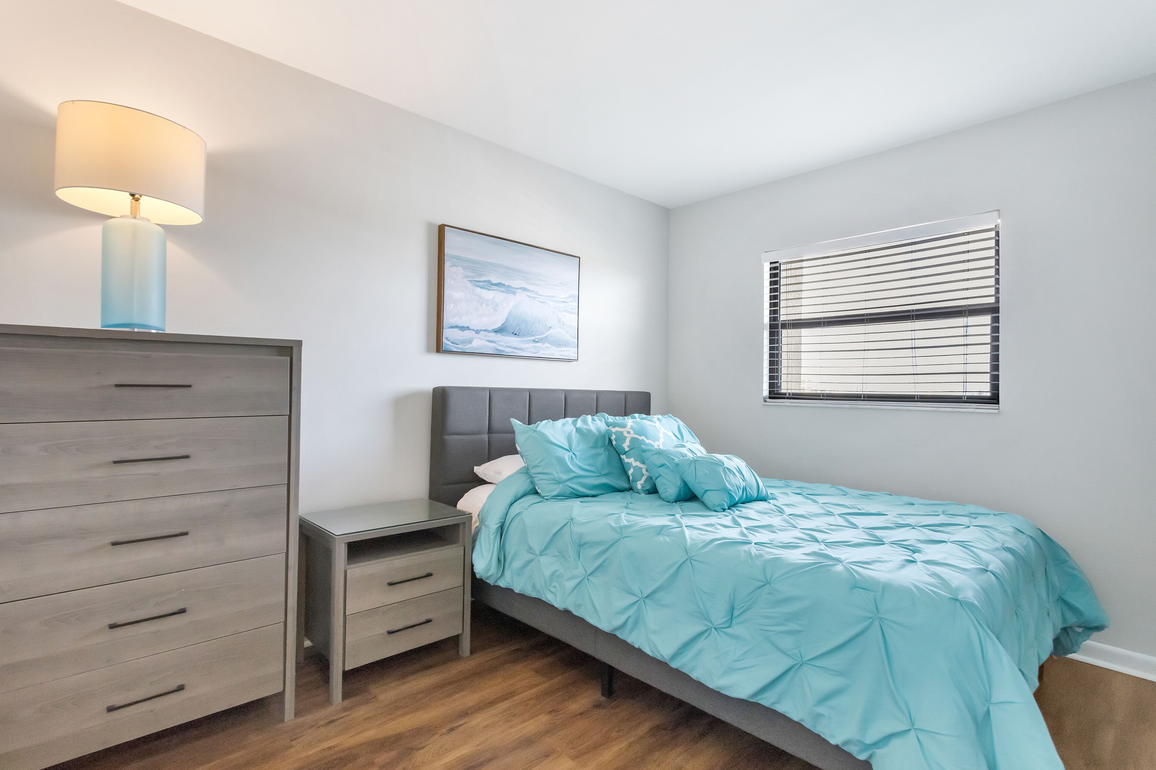 A bedroom with a bed, a dresser, a nightstand, a lamp, a window with blinds, and a picture on the wall.