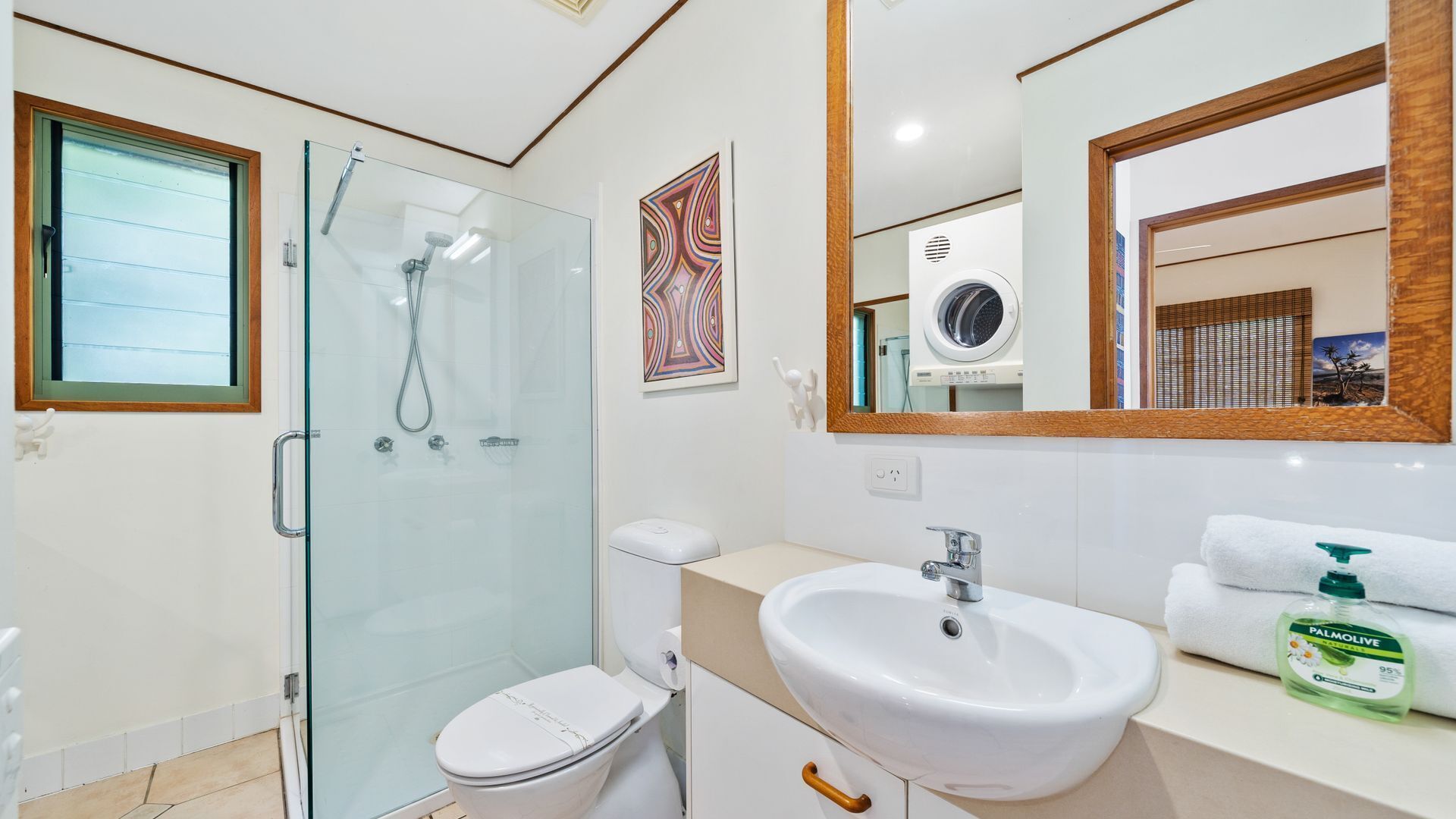 A modern bathroom with a glass shower enclosure, a toilet, a sink with a mirror above, and a towel dispenser.