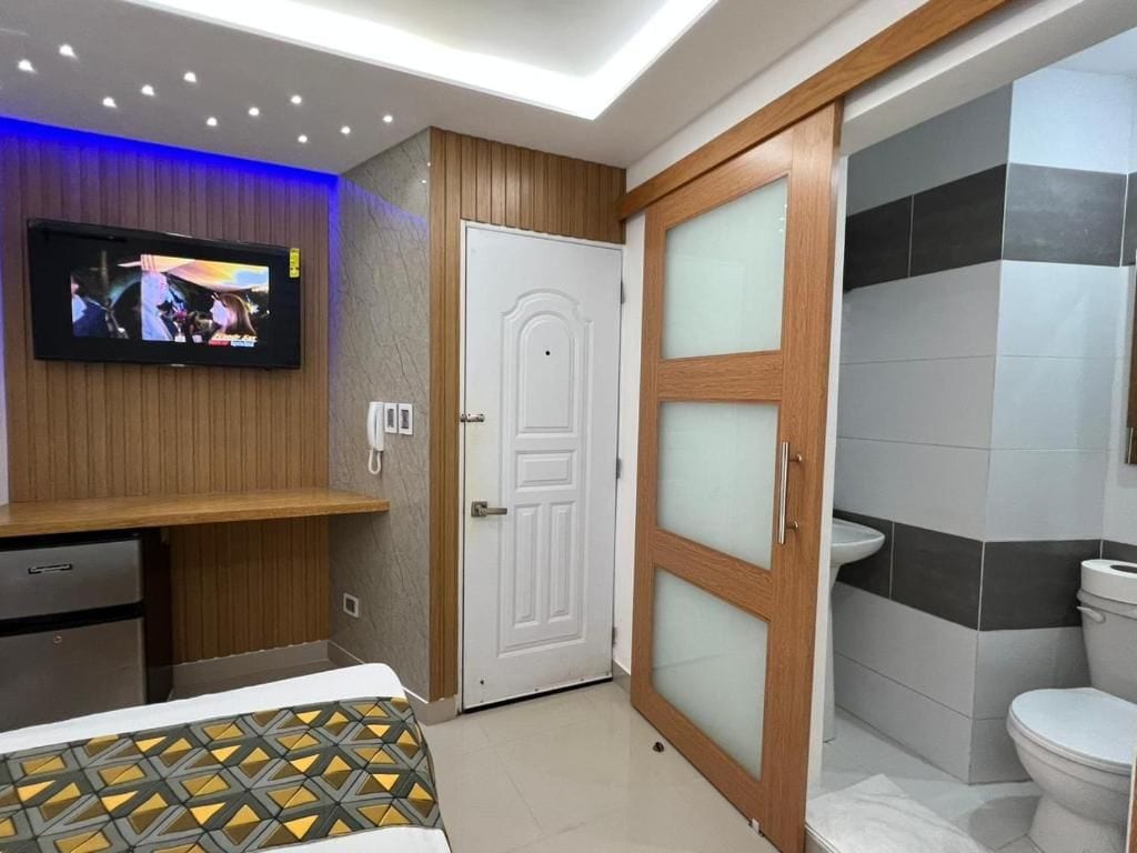 A hotel room with a television mounted on the wall, a wooden cabinet, a bed with a patterned bedspread, a white door, and a bathroom with a toilet and sink.