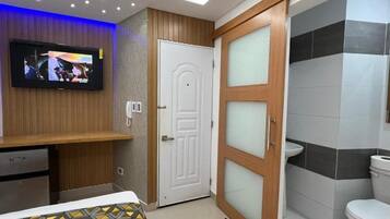 A hotel room with a television mounted on the wall, a wooden cabinet, a bed with a patterned bedspread, a white door, and a bathroom with a toilet and sink.