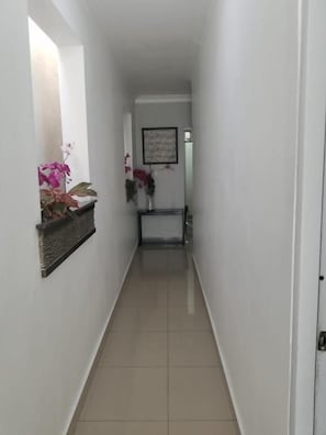 A narrow hallway with a tiled floor, a door on the right, and a small table with a vase of flowers on the left. - Alegría Hostal (Santiago de los Caballeros)