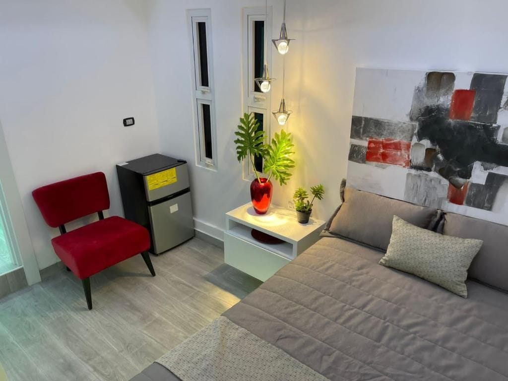 A bedroom with a bed, a red chair, a desk, a plant, and a painting on the wall.