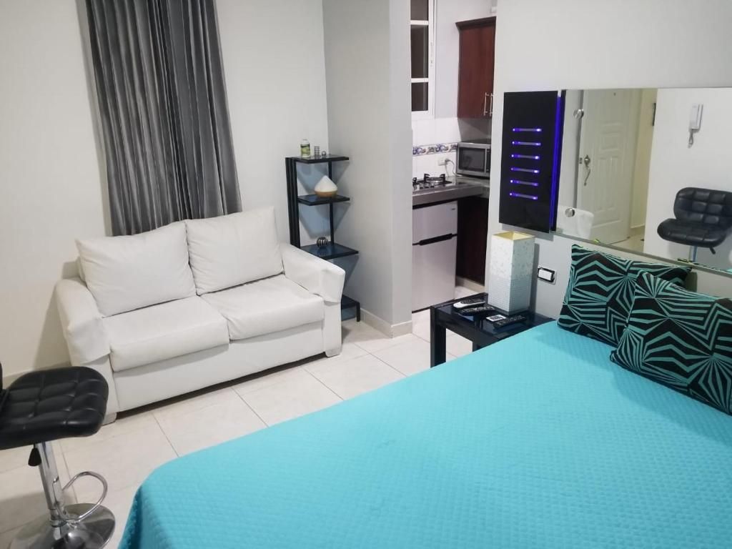 A bedroom with a bed, a white sofa, a small table, a mirror, and a kitchenette in the background.