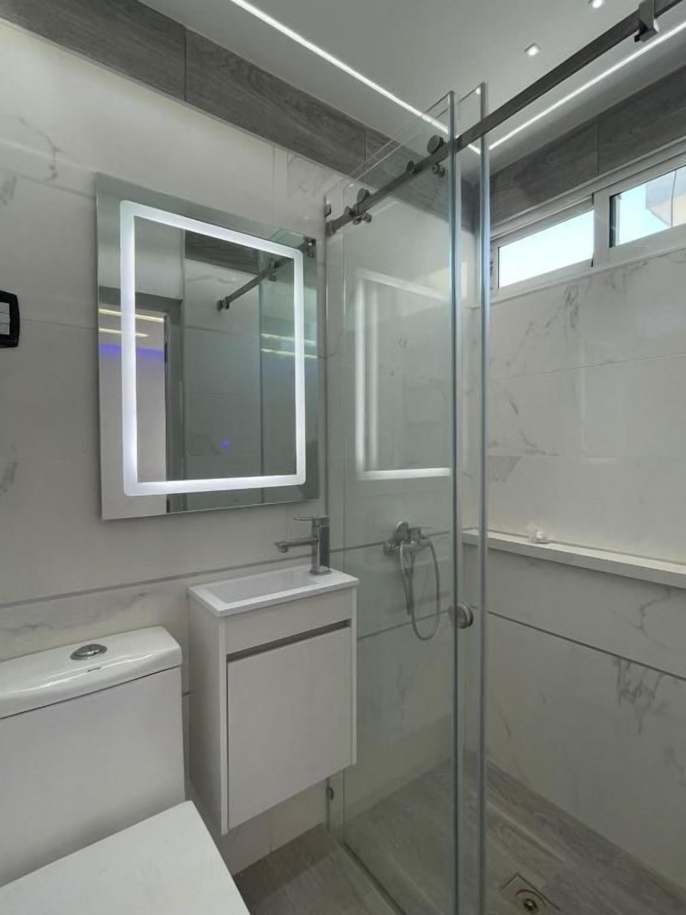 Un baño moderno con cabina de ducha de vidrio, lavabo blanco e iluminación integrada en el espejo.