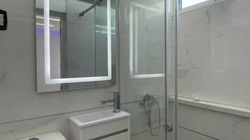 Un baño moderno con cabina de ducha de vidrio, lavabo blanco e iluminación integrada en el espejo.