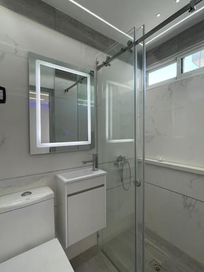 A modern bathroom with a glass shower enclosure, a white sink, and a mirror with integrated lighting. - Alegría Hostal (Santiago de los Caballeros)