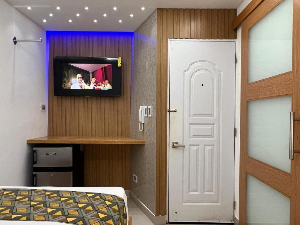 A room with a television mounted on the wall, a wooden cabinet, a white door, and a bed with a patterned bedspread.