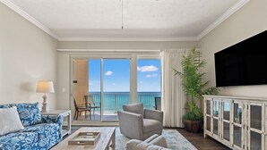 A living room with a sofa, armchairs, a coffee table, a TV, and a view of the ocean through large sliding doors. - Treasure Island 0403 (Panama City Beach)