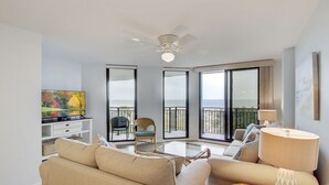 Een ruime woonkamer met een bank, een salontafel en een televisie op een standaard. Grote ramen bieden uitzicht op de zee.