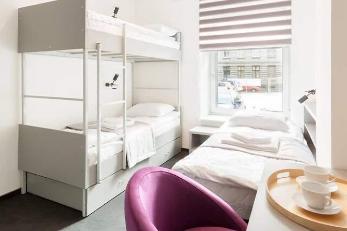 A modern hotel room with a bunk bed, a desk with a tray of cups, a purple chair, and a window with blinds.
