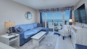 Un espace de vie comprenant un canapé, une table basse, une lampe, un fauteuil et une vue sur la mer depuis un balcon.