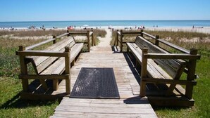 Une promenade en bois mène à une plage où se trouvent des bancs et un tapis noir au centre.