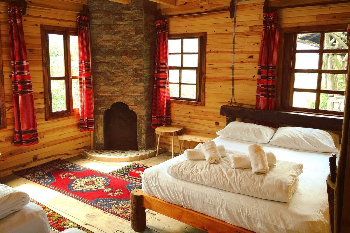 A wooden cabin room with a stone fireplace, a bed with white linens, and red curtains.