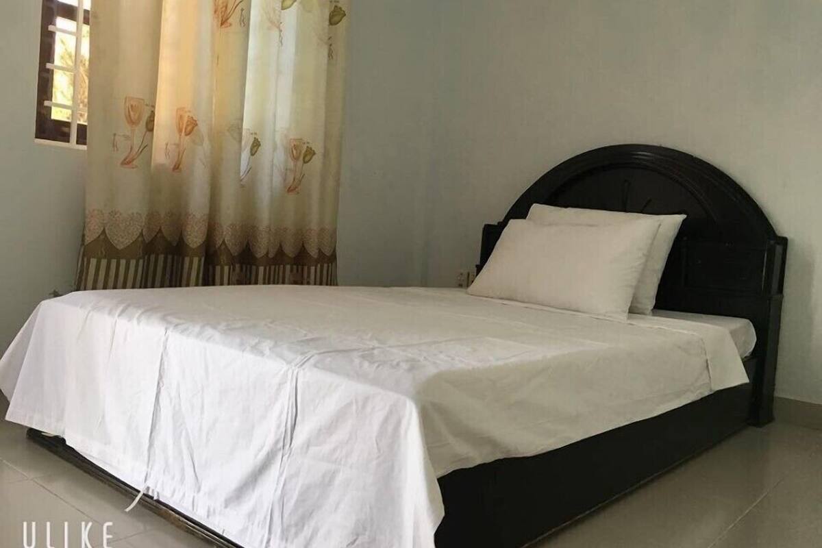 A bed with a dark headboard and white bedding in a room with a plain wall and a window with curtains.