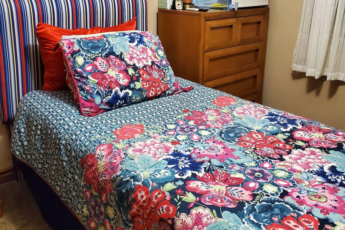A bedroom with a bed, a dresser, a lamp, a framed picture, and a refrigerator.