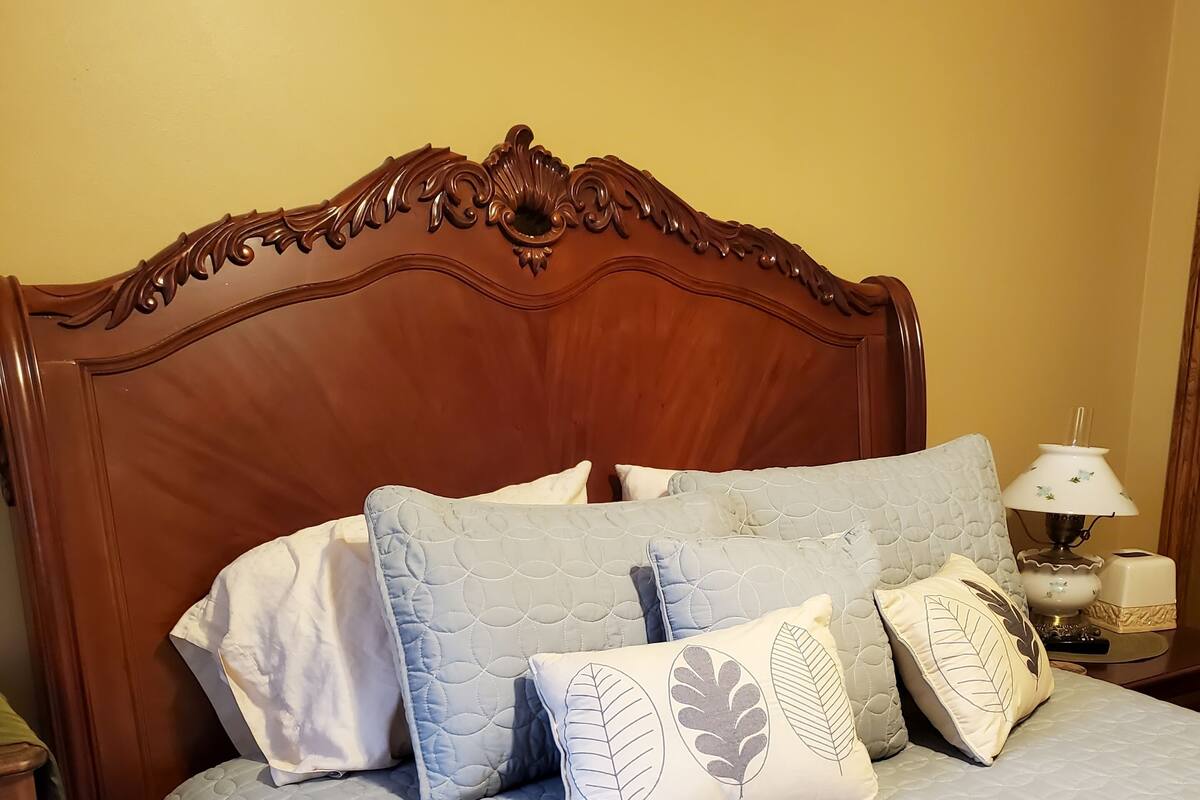 A neatly made bed with a quilted blue cover and decorative pillows, a wooden headboard, and a bedside table with a lamp and a box.