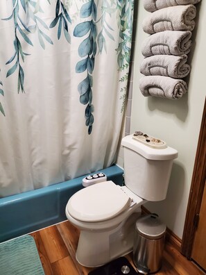 Un baño con inodoro, bidé, un cubo de basura, toallero con toallas dobladas y cortina de ducha con diseño de hojas.