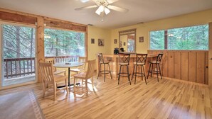 Ein Zimmer mit Holzboden, einem Esstisch und Stühlen, einer Bar mit Hockern, großen Fenstern mit Blick auf einen Wald und einem Deckenventilator.