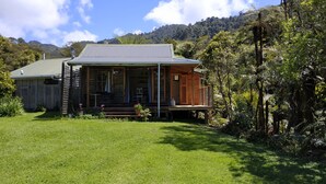 A wooden house with a porch, surrounded by lush greenery and a mountain backdrop. - Opua Garden Cottage, sleeps 3 (Collingwood)