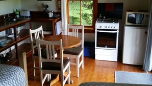 A compact kitchen with a stove, microwave, and sink. There is a dining table with chairs and a window with a view of greenery. - Opua Garden Cottage, sleeps 3 (Collingwood)