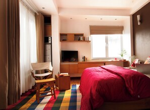 A bedroom with a bed, a desk, a chair, a TV, and a window with blinds.