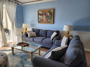 Uma sala de estar com um sofá azul, duas luminárias, um quadro e uma mesa de centro de vidro.