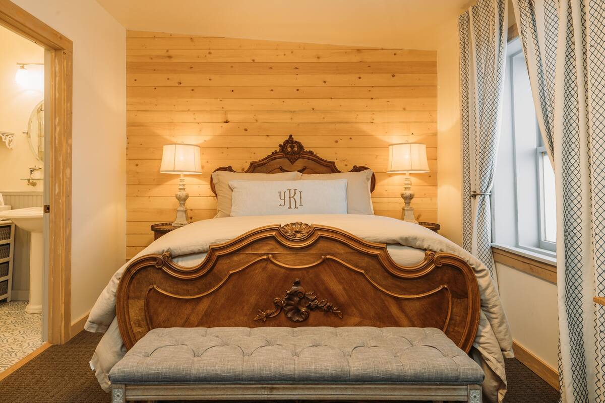 A bedroom with a wooden headboard, two bedside lamps, a bench, and a window with patterned curtains.