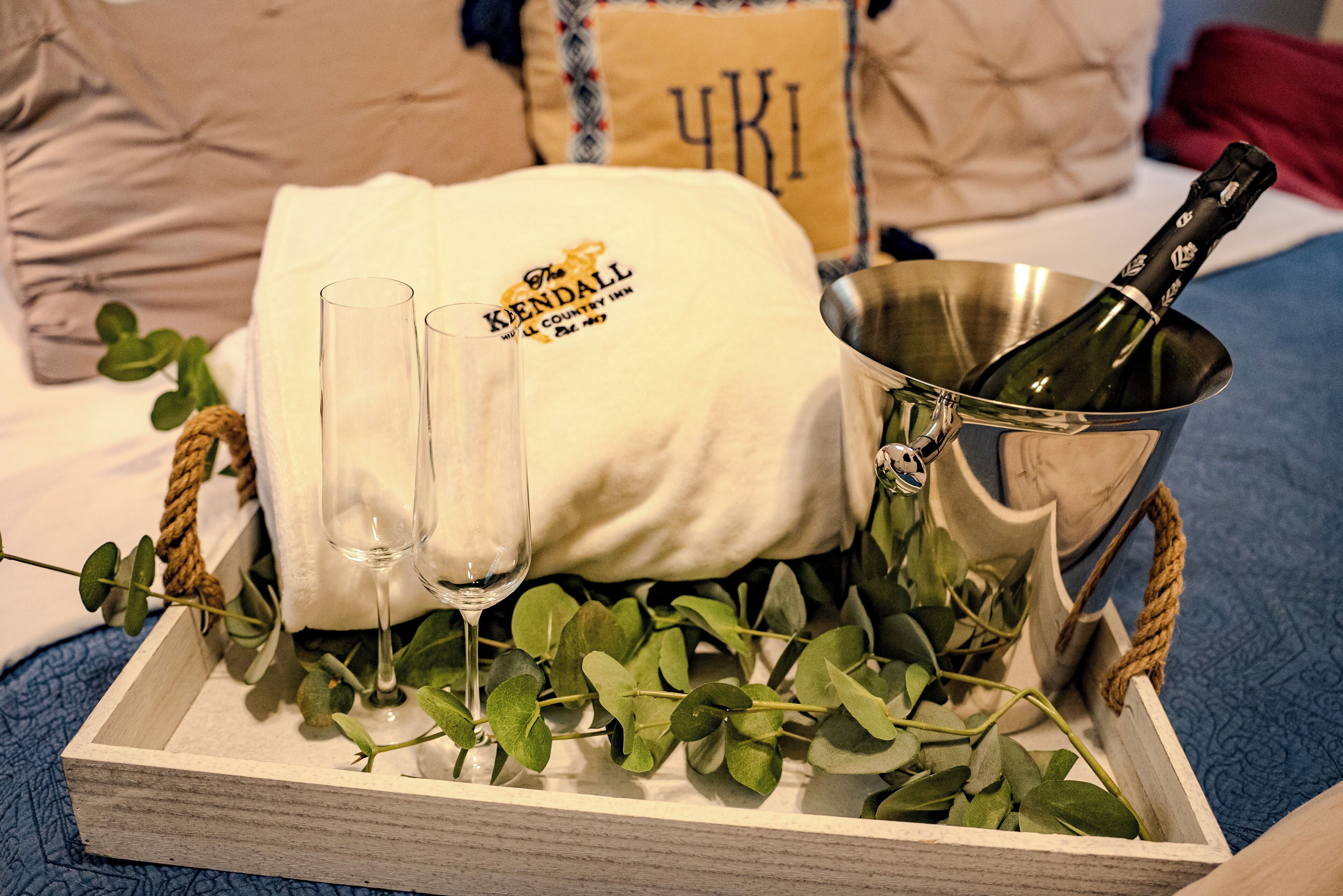 Una bandeja de madera con una botella de champán, dos copas y una toalla blanca con un logotipo, sobre una cama con una colcha azul y un almohadón con el logo '4K'.