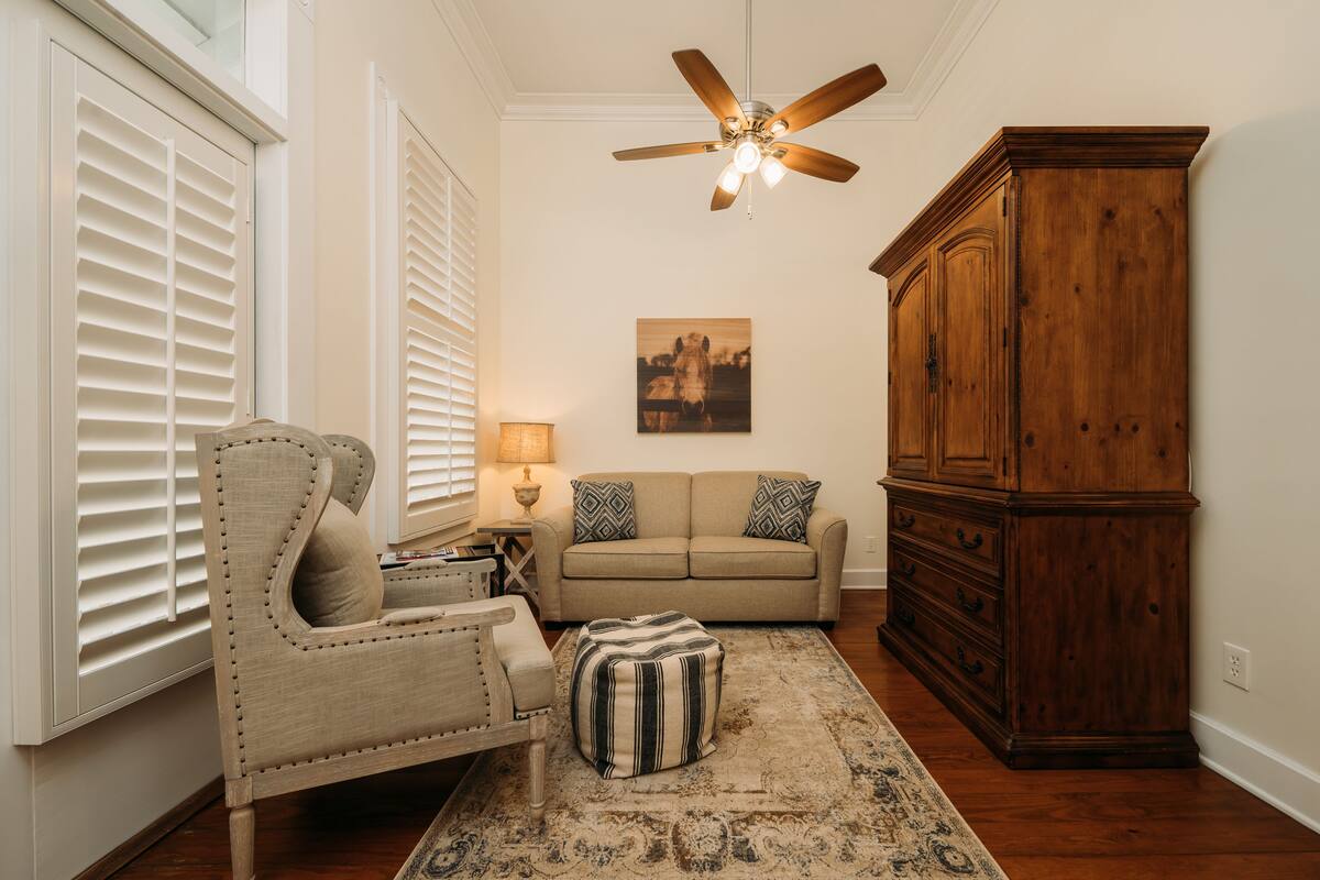 A living room with a beige sofa, a wooden cabinet, a striped ottoman, and a ceiling fan.