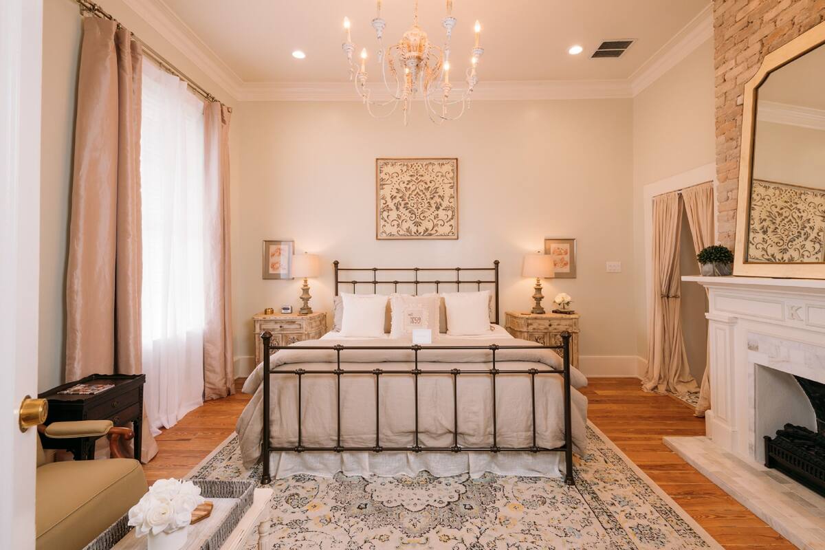 A bedroom with a bed, bedside tables, a chandelier, a fireplace, and a large mirror.