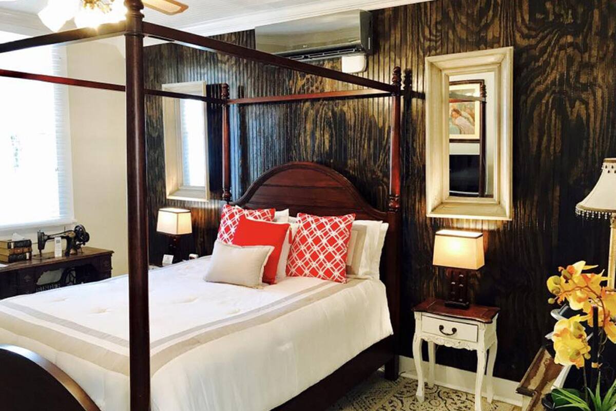 A four-poster bed with a wooden frame, white bedding, and red and white patterned pillows. There is a nightstand with a lamp and a small table with a lamp beside the bed. A mirror hangs on the wall above the bed.