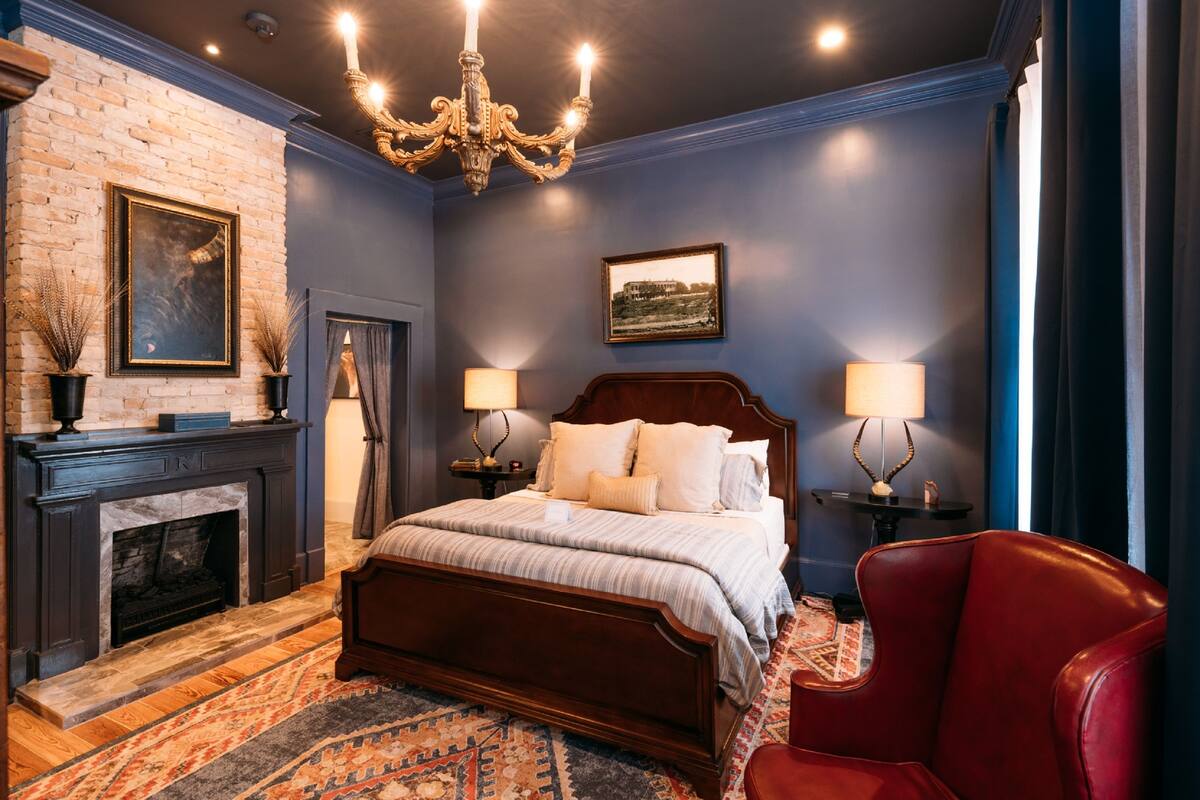 A bedroom with a large bed, a fireplace, a chandelier, and a red armchair.