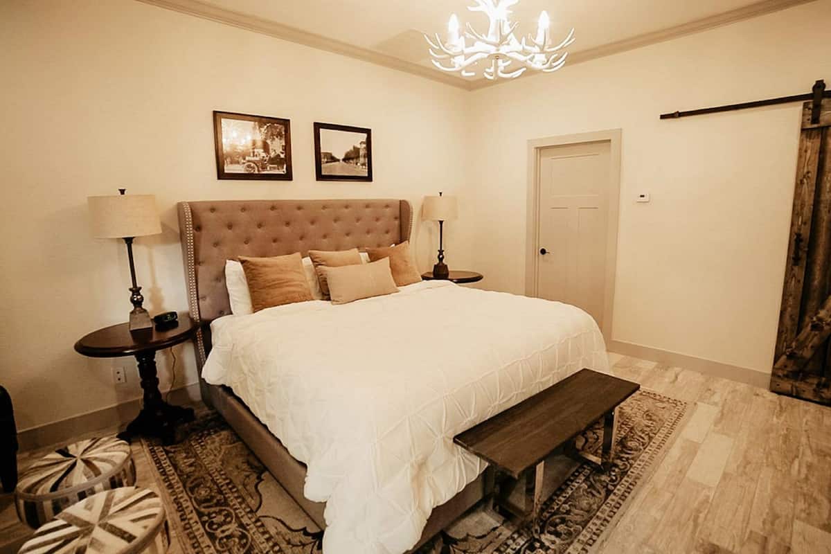 A bedroom with a large bed, two bedside tables, a bench, a chandelier, and a door.