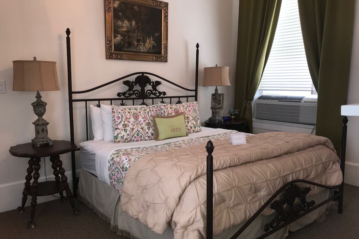 A neatly made bed with a quilted cover, two bedside tables with lamps, a framed painting on the wall, and a window with curtains.