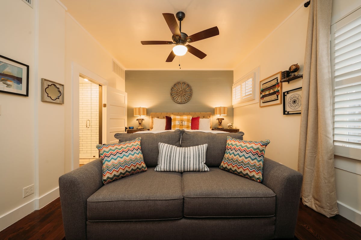 A gray sofa with patterned pillows, a ceiling fan, and wall-mounted decor in a well-lit room with hardwood flooring.