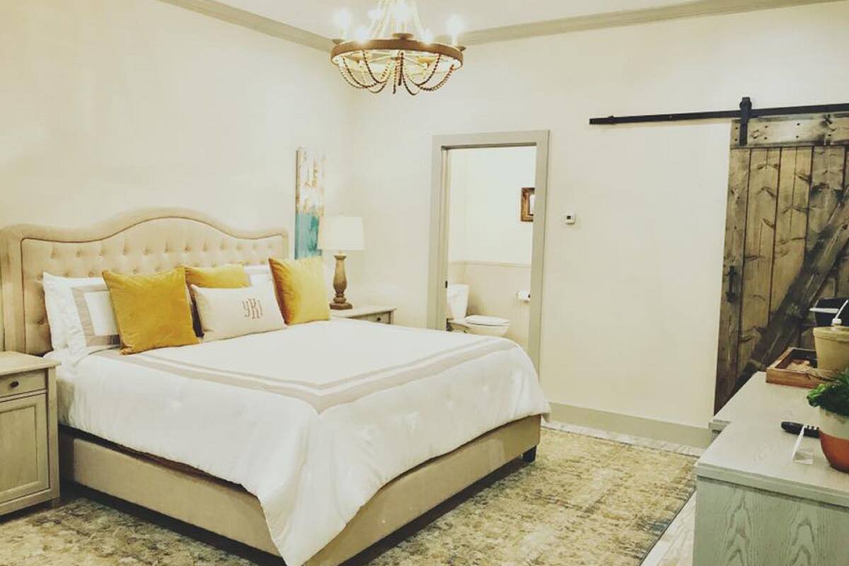 A bedroom with a large bed, a chandelier, a nightstand, a dresser, and a door with a wooden door frame.