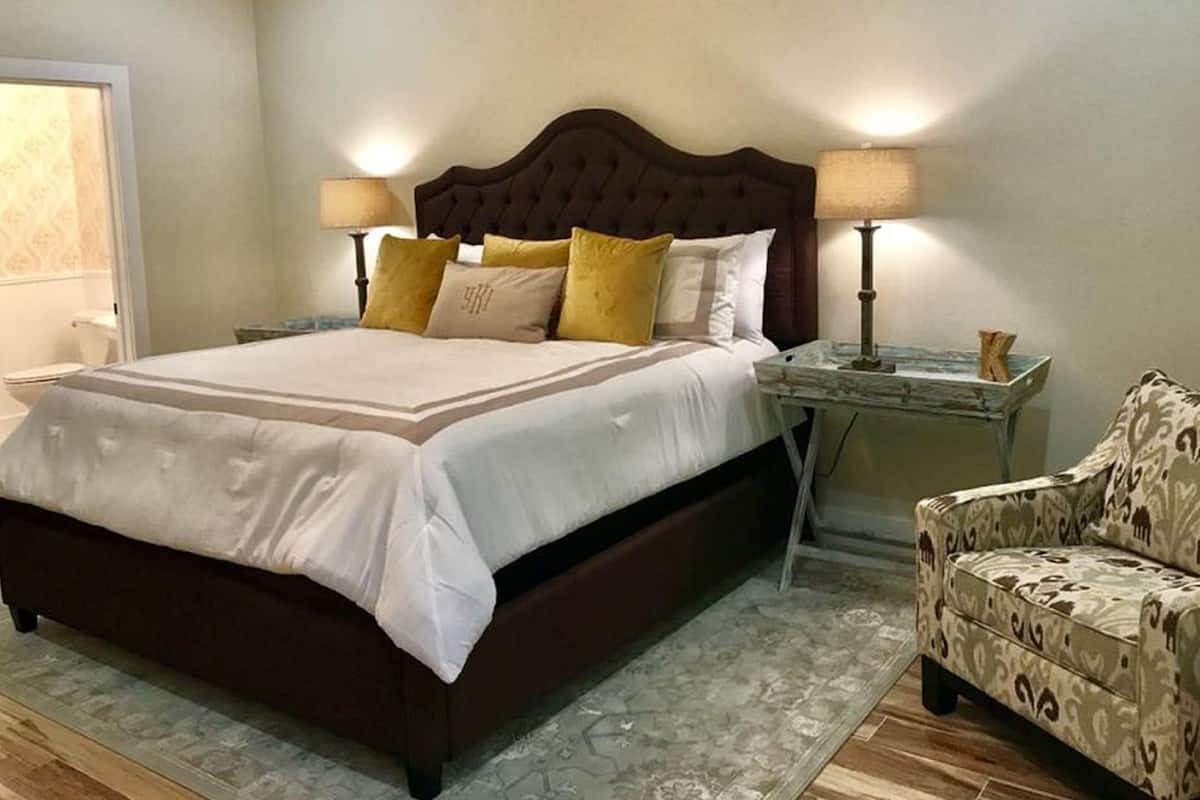 A neatly made bed with a dark headboard, two bedside lamps, and a patterned armchair.