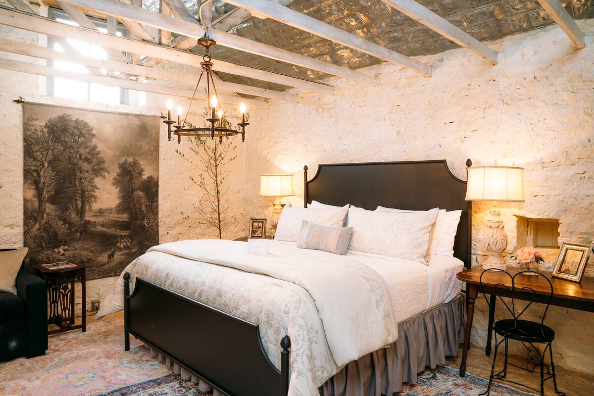 A bedroom with a large bed, two bedside tables with lamps, a chandelier, and a large landscape painting on the wall.