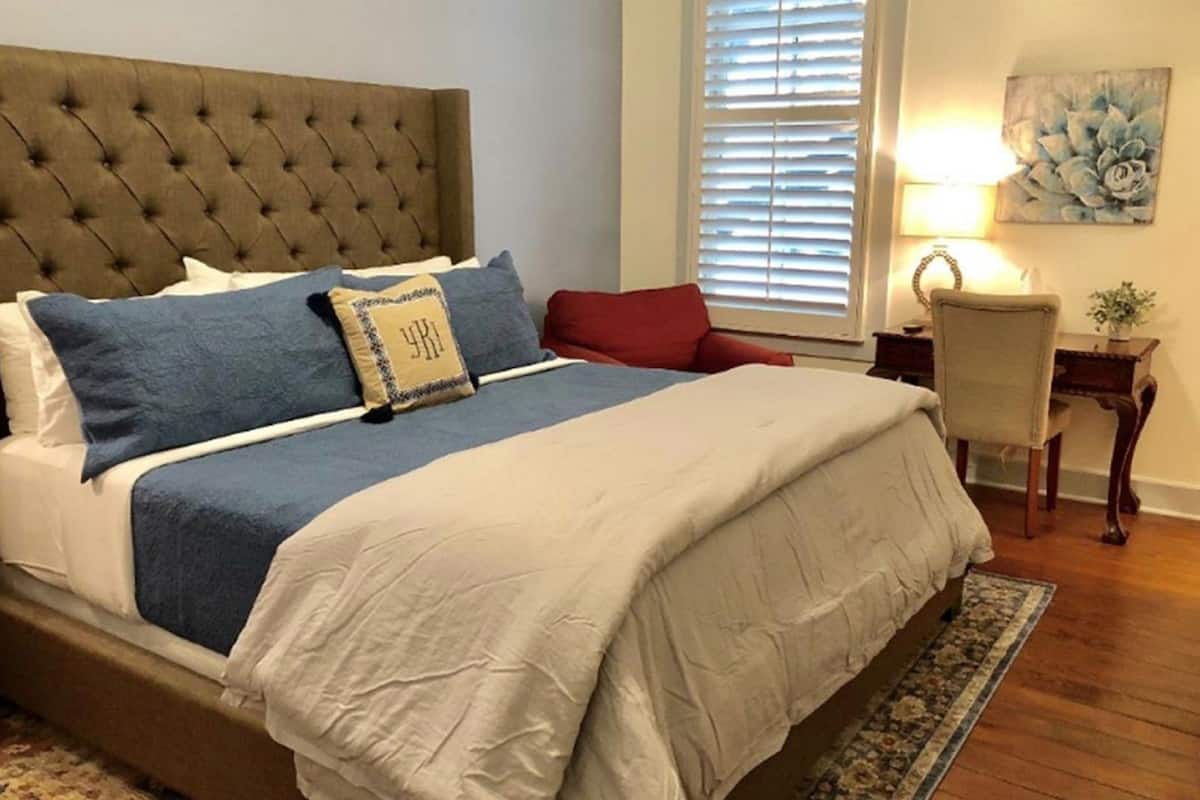 A bedroom with a tufted headboard, a large bed with white and blue bedding, a red pillow, a side table with a lamp, and a painting on the wall.