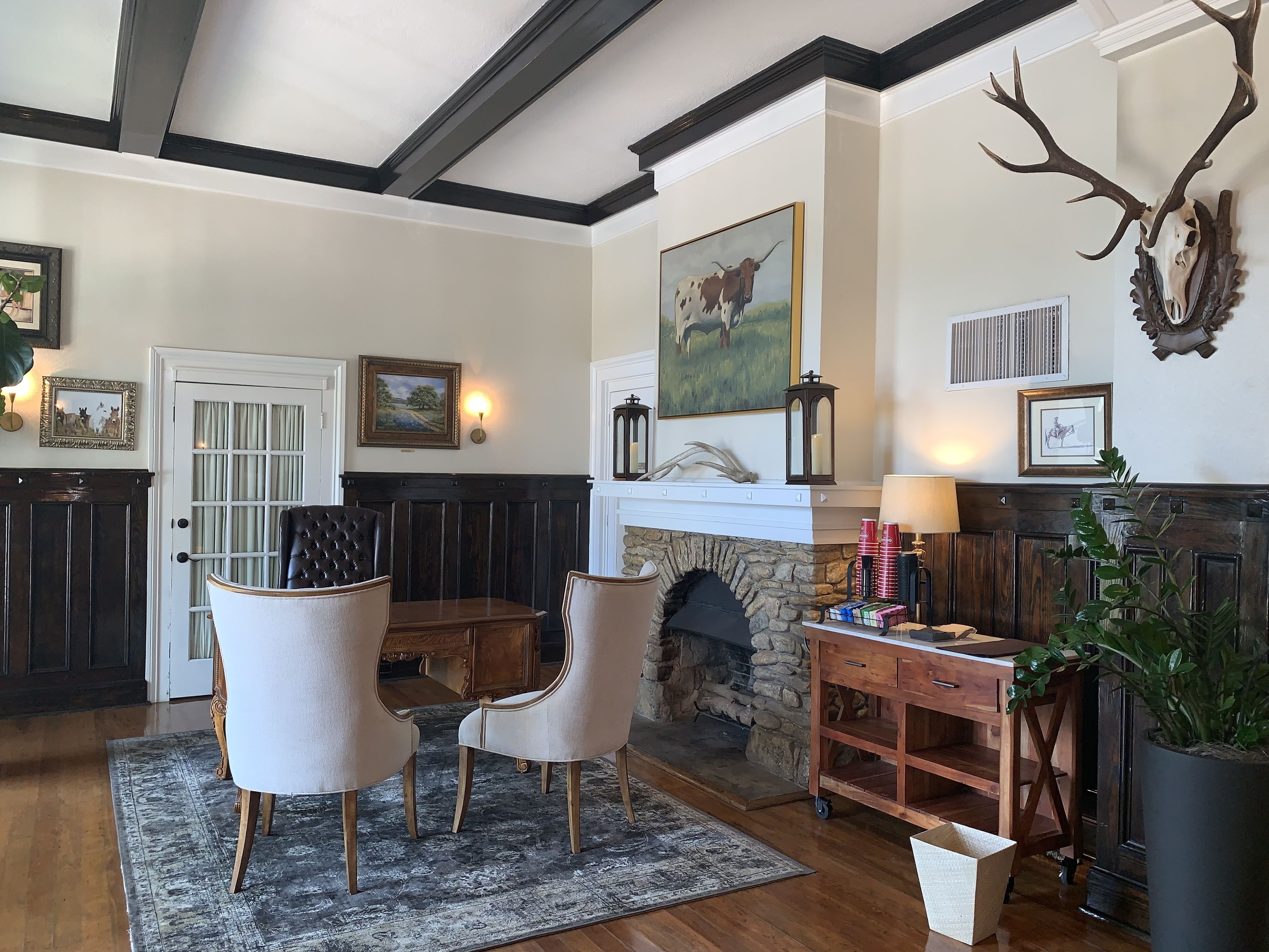 Un salon chaleureux avec une cheminée en pierre, un coin repas avec une table et des chaises, et un grand crâne de cerf accroché au mur.