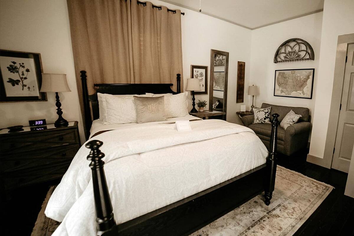 A neatly arranged bedroom with a large bed, a bedside table, a nightstand, a chair, and a door.