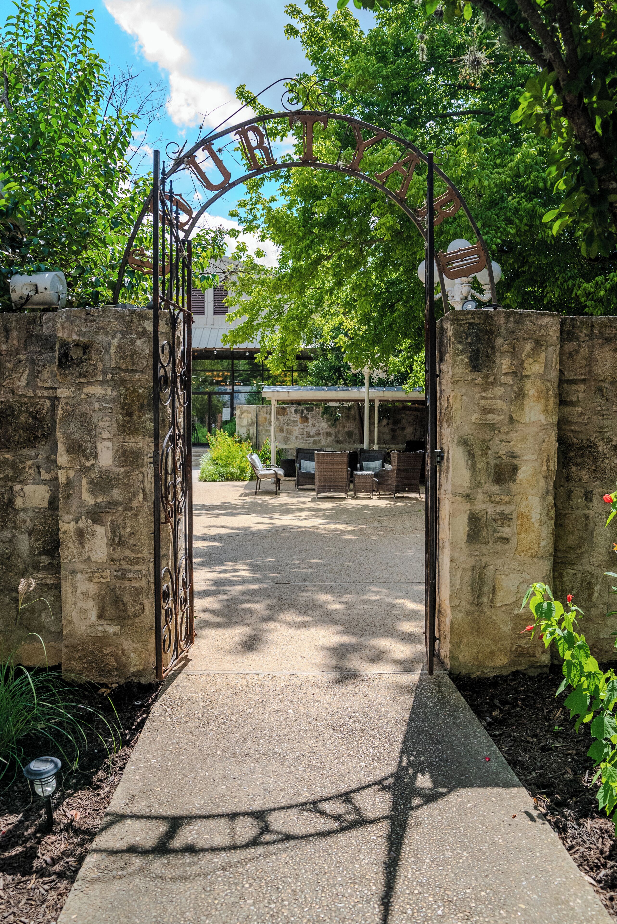 A wrought iron gate with the word "TRINITY" arched above it, leading to a courtyard with stone walls, outdoor seating, and greenery.