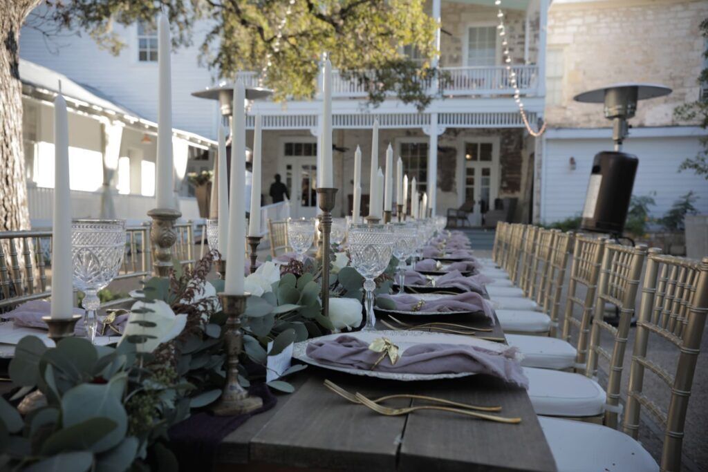 An outdoor dining area with tables set for a meal, surrounded by white columns and a balcony in the background.