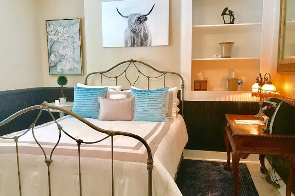 A hotel room with a metal bed frame, a blue and white striped pillow, a white pillow, a small potted plant, a framed picture of a bull, a shelf with decorative items, and a wooden chair.