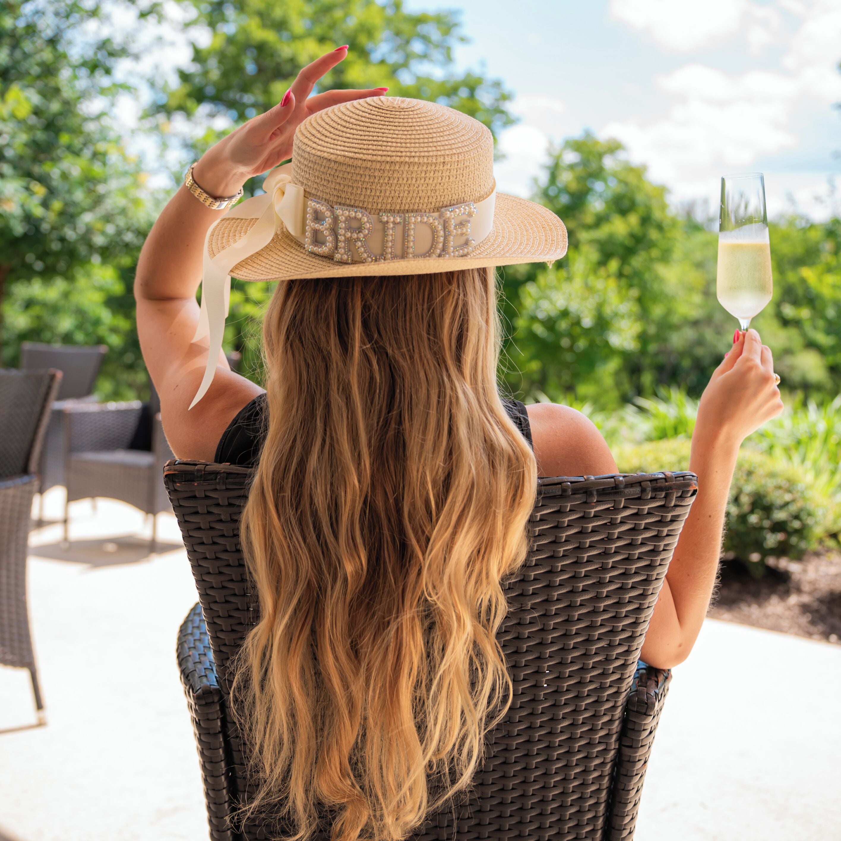 Une personne assise sur une chaise en osier à l’extérieur, tenant un verre de champagne, portant un chapeau de paille sur lequel est écrit le mot « MARIÉE », avec un décor luxuriant et verdoyant en arrière-plan.