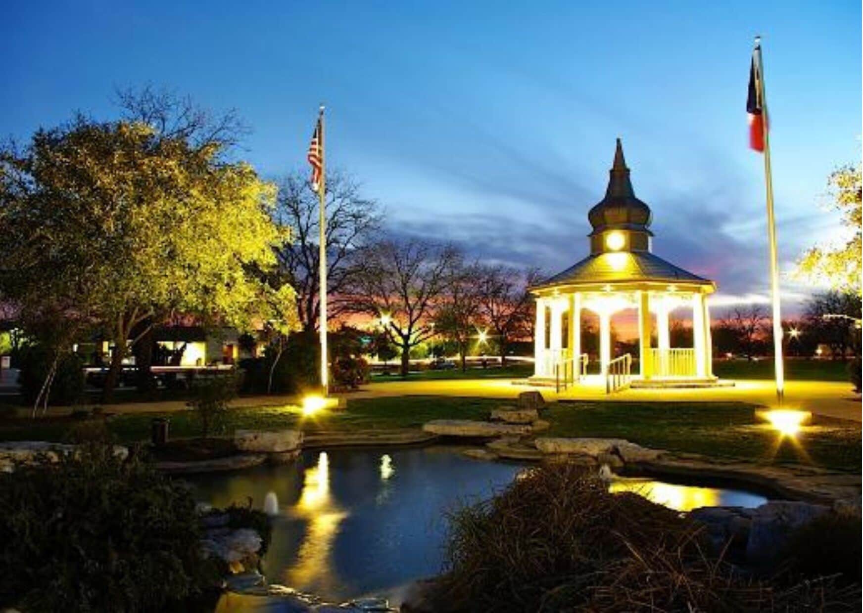 A gazebo with a dome roof and a small pond in front, surrounded by trees and flags.