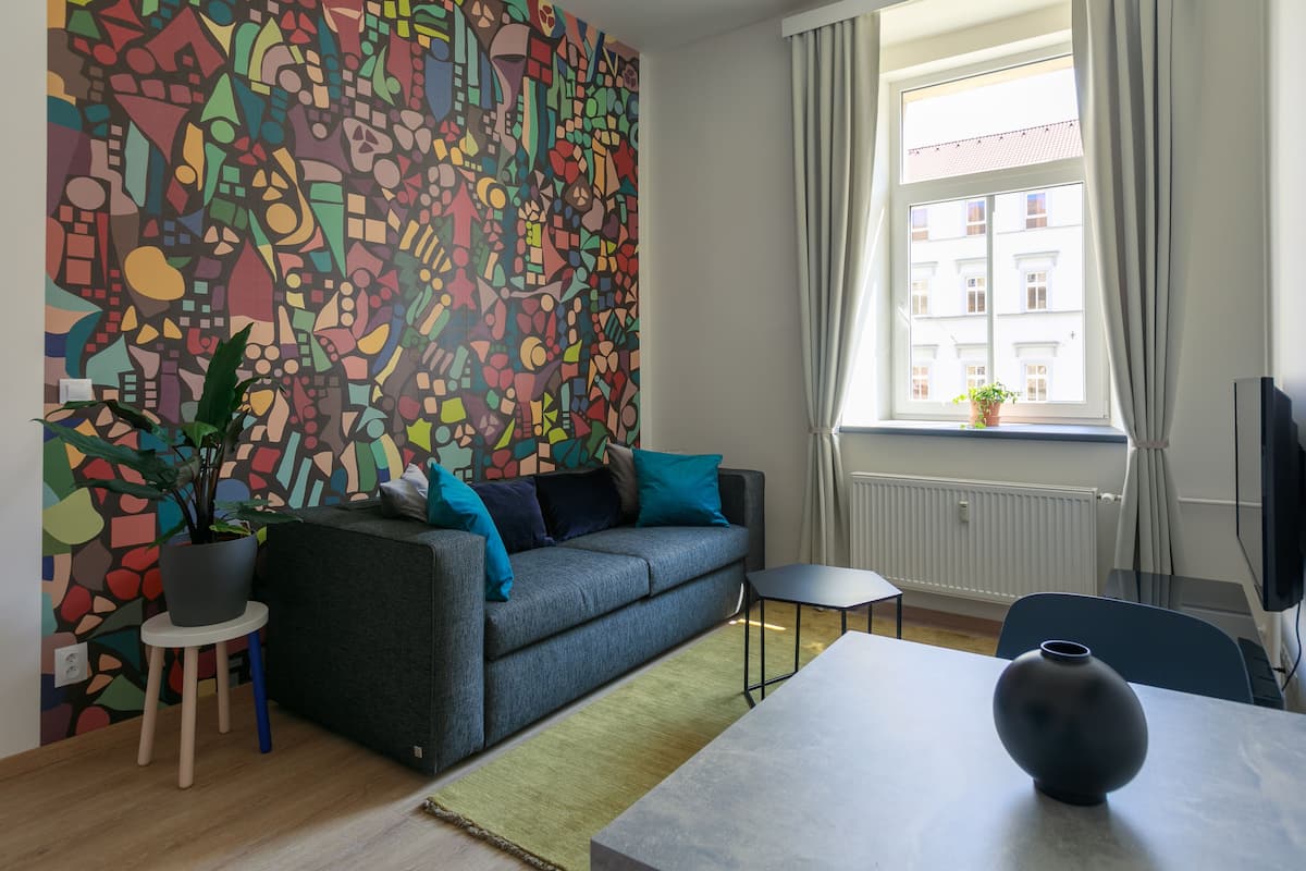 A modern living room with a colorful abstract wall, a grey sofa with teal cushions, a small round table, and a potted plant.