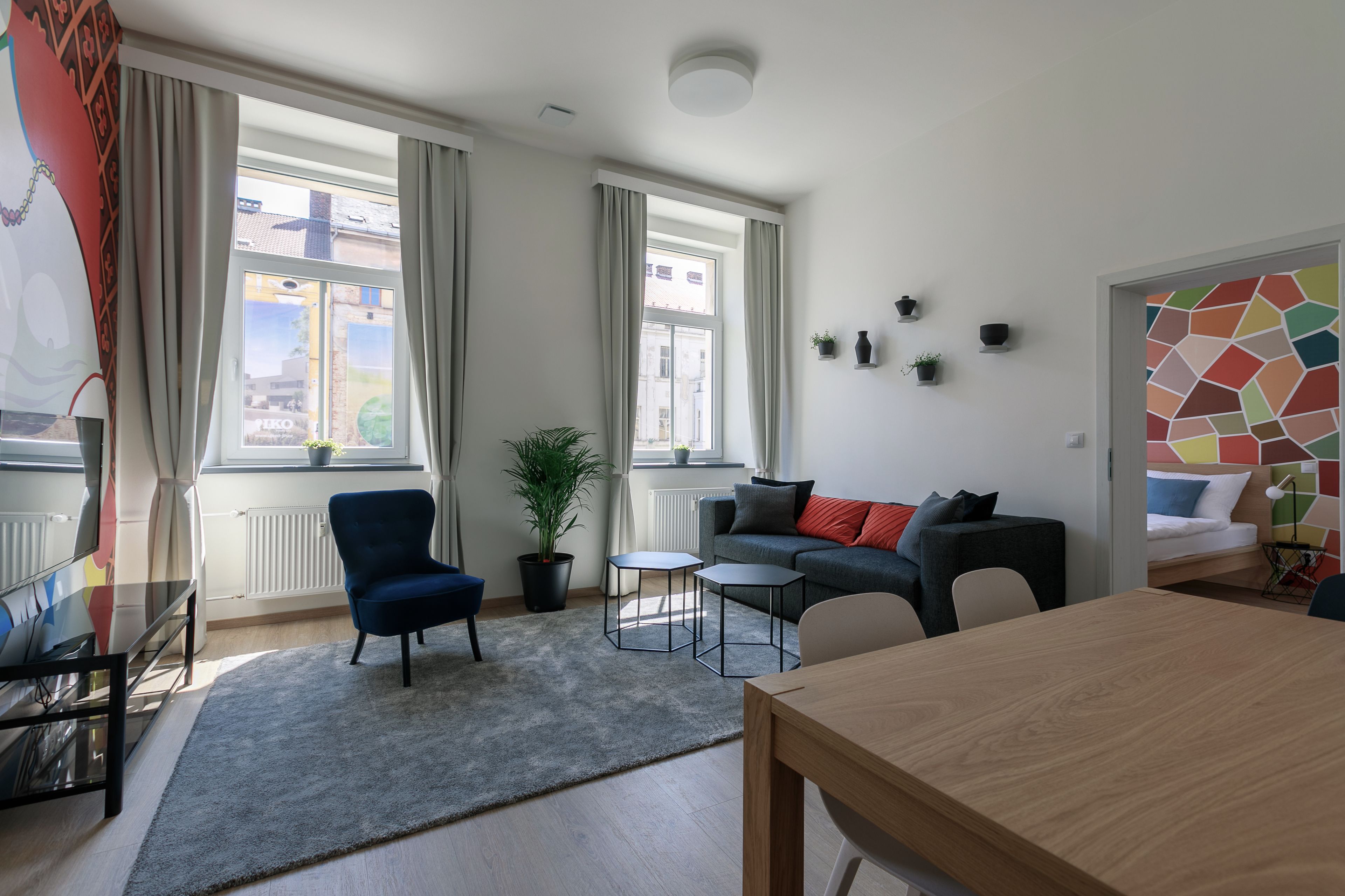 A modern living room with a sofa, a dining table, and a blue armchair. There are windows with curtains, a potted plant, and a colorful wall art piece.